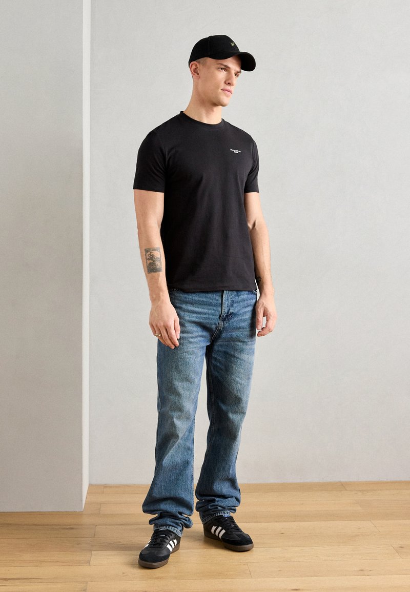 Tricou negru din bumbac cu guler rotund, asortat cu blugi din denim bleu deschis și tenisi negri cu dungi albe. Se poartă o șapcă neagră.