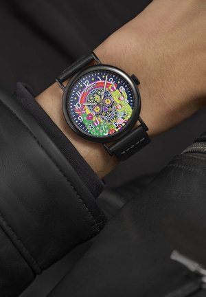 Montre Timex colorée présentant un design de crâne sur le cadran, avec un bracelet en cuir noir et un boîtier rond. Des détails floraux et des accents vibrants sont présents.