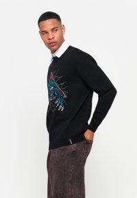 Man met een zwart sweater met een brullende blauwe tijgerafbeelding over een witte colbert en kastanjebruine gewassen spijkerbroek, kijkend opzij met een hand in zijn zak.