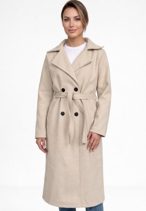 Kvinde iført en lang beige dobbeltradet frakke med bælte, stående mod en ensartet hvid baggrund.