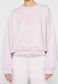 Sweatshirt rose clair avec un motif texturé en cercle et barre à l'avant. Présente des manches larges et un ourlet côtelé. Associé à un pantalon à motifs.