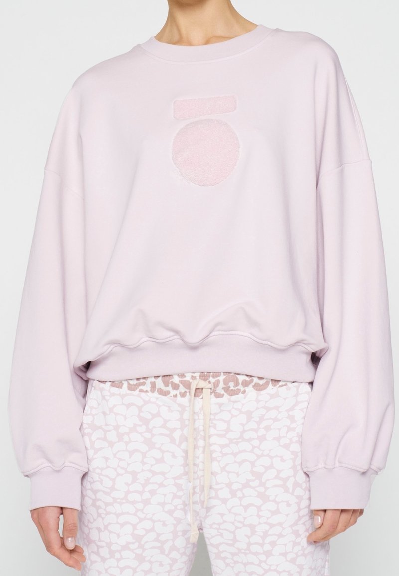 Sweatshirt rose clair avec un motif texturé en cercle et barre à l'avant. Présente des manches larges et un ourlet côtelé. Associé à un pantalon à motifs.