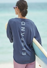 Marineblaues Langarmshirt mit dem Wort "ELBSAND" vertikal in hellgrau bedruckt. Kombiniert mit rosa Hose, hält ein Surfbrett.