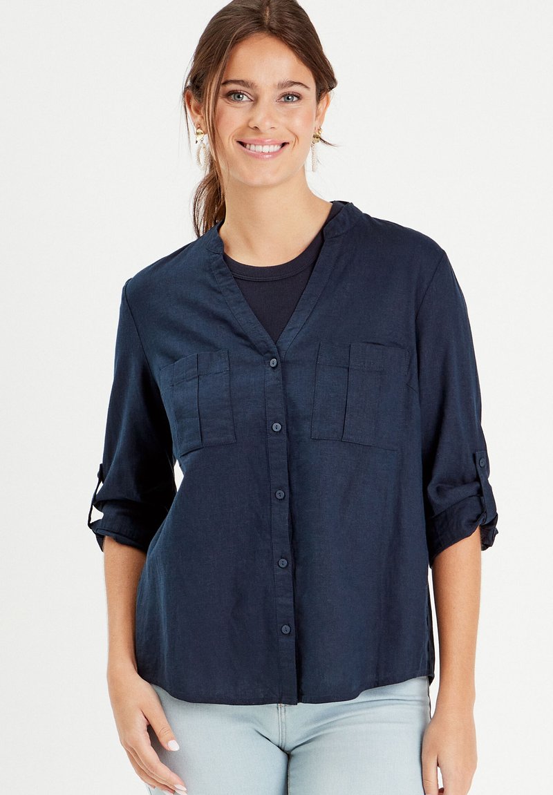 Chemise en lin bleu marine avec un col en V, deux poches poitrine et des manches retroussées. Texture douce et détail de fermeture à boutons.