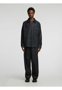 Tumšas džinsa jaka ar pogām priekšpusē un krūšu kabatu, valkāta kopā ar vaļīgām tumšām biksēm un melniem apaviem. Gluda tekstūra.