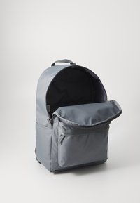 Mochila de tecido cinza com um topo arredondado, fecho de zíper, bolso frontal e um interior preto contrastante. Textura suave e design simples.