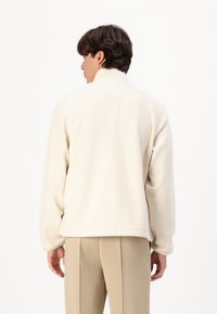 Cream fleece jacket with a high collar, loose fit, and ribbed hem. Soft texture and no visible logos or hardware details.