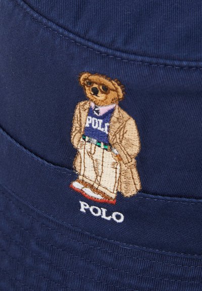 Navy plavi šešir s širokim obodom sadrži izvezenu medu s bež kaputom preko plave majice s logotipom "POLO", bijelim cipelama i vidljivim detaljima šavova.