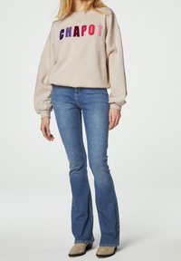 Felpa beige con lettere ricamate multicolori "CHAPOT" sulla parte anteriore, abbinata a jeans blu attillati a zampa d'elefante e calzature di colore chiaro.