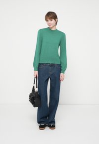 Giovane in maglione verde e jeans blu larghi, con una borsa nera in mano, in piedi davanti a uno sfondo bianco uniforme.