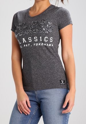 Kobieta w dopasowanym, ciemnoszarym t-shircie z cekinowym logo i białym napisem "Classics, Yokohama", w zestawie z jasnoniebieskimi jeansami.