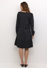 Zwarte jurk met lange mouwen, geplooide zoom en een vierkante halslijn. Beschikt over een ritssluiting aan de achterkant en iets geplooide mouwdetails. Glad stof.