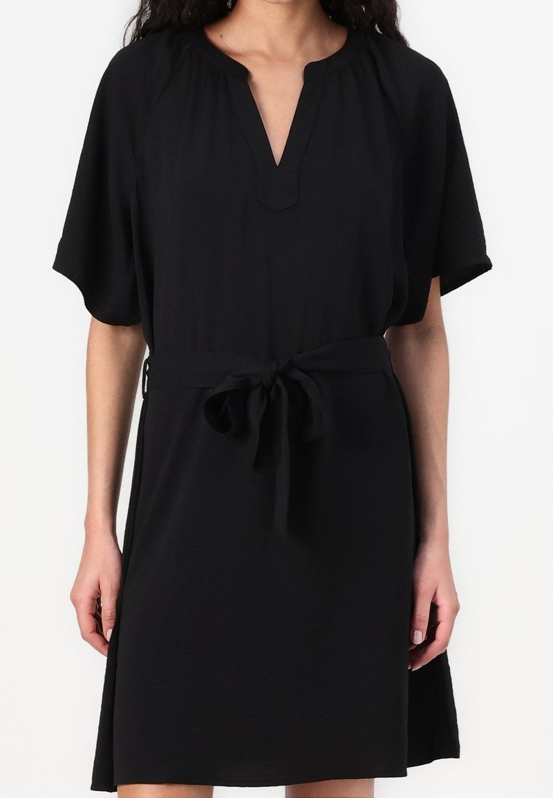 Femme portant une robe noire mi-longue à manches courtes, avec un col en V et une ceinture nouée à la taille sur un fond uni.