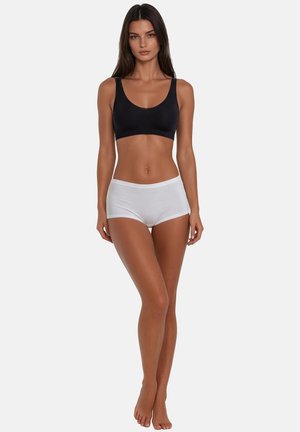 Donna in piedi a piedi nudi, rivolta in avanti, che indossa un reggiseno sportivo nero e mutandine bianche modello boyshort su uno sfondo chiaro uniforme.