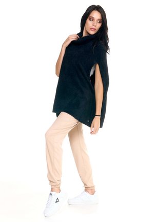 Une femme porte un haut noir sans manches à col bénitier, un pantalon de jogging beige et des baskets blanches sur fond blanc.