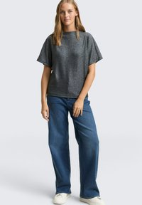 Lesklý šedý top s krátkymi rukávmi a voľným strihom, kombinovaný s modrými nohavicami so širokými nohavicami a bielymi teniskami na neutrálnom pozadí.