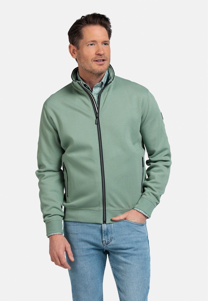 Lichtgroene zip-up jack van een zachte stof met zwarte ritsaccenten, voorzien van zijzakken en geribde mouwen, gedragen over een bedrukt shirt.