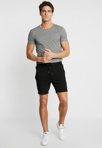 Graues Rundhals-T-Shirt im körpernahen Schnitt, kombiniert mit schwarzen Shorts, elastischem Bund mit Zugband. Das Model trägt weiße Sportschuhe.