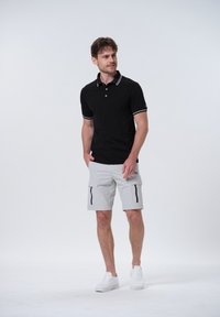 Polo negro con acentos blancos, combinado con pantalones cortos cargo grises claros con cremalleras laterales y zapatillas blancas. Diseño minimalista en general.