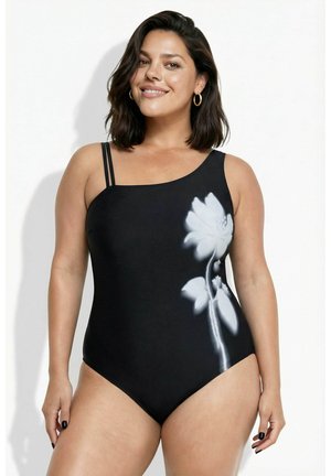 Femme portant un maillot de bain noir à une épaule avec de fines bretelles doubles et un grand motif floral blanc sur le côté, souriante sur un fond blanc.