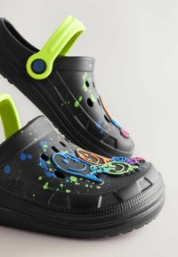 Schwarze Kunststoff-Clogs mit leuchtend grünen Riemen und bunten Smiley-Gesichtsverzierungen, ergänzt durch blau-grüne Spritzmuster.