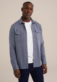 Blauwe button-up shirt voor heren met twee borstzakken, lange mouwen en een zachte textuur, gedragen over een wit T-shirt, gecombineerd met donkere jeans.