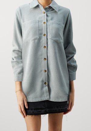Person trägt ein hellblaues, geripptes Hemd mit Knöpfen und zwei Brusttaschen sowie einen schwarzen, ausgefransten Jeansrock.