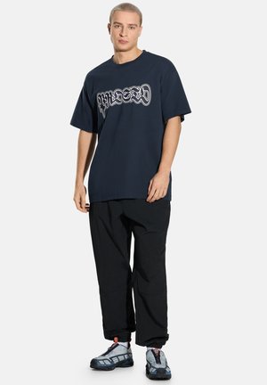 Granatowy, oversize'owy t-shirt z białym nadrukiem, zestawiony z czarnymi spodniami i szarymi butami sportowymi z czarnymi i czerwonymi akcentami.