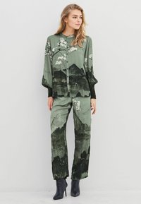 Langærmet grøn bluse og matchende bukser med et bjerg- og blomstermønster. Blødt stof, knaplukning, løs pasform, vidde ben.