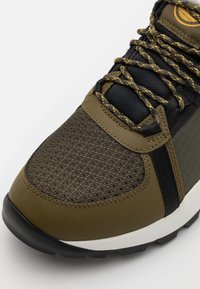 Zapatilla verde oliva con malla texturizada y paneles sólidos, cordones negros y una suela negra resistente. Presenta un cuello acolchado y detalle de marca.