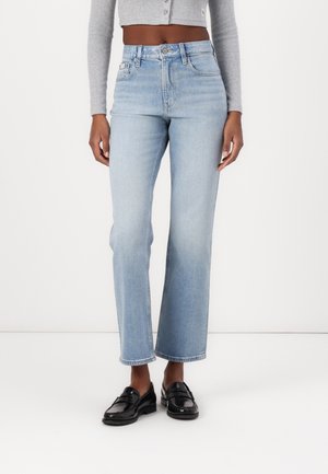 Osoba ubrana w jasnoniebieskie, wysokowatowe jeansy o prostych nogawkach, szary krótki top z długim rękawem oraz czarne mokasyny, stojąca na tle gładkiej białej ściany.