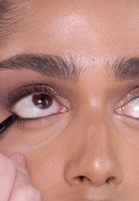 Zwarte eyeliner is aangebracht op het onderste ooglid. De ogen hebben een warme bruine tint, met goed gedefinieerde wimpers en een gladde, egale teint.