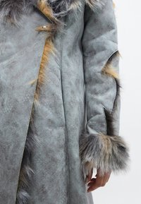 Gaišgriša mākslīgā zamšādas mētelis ar teksturētu apdari, ar tumšgrišiem un smilšu krāsas mākslīgā zamšādas akcentiem uz piedurknēm un apkakles, kā arī pogu aizdariem.