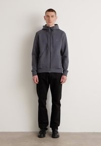 Sudadera gris con cierre y capucha con cordón, textura suave y un bolsillo frontal, combinada con pantalones negros y zapatos negros resistentes.