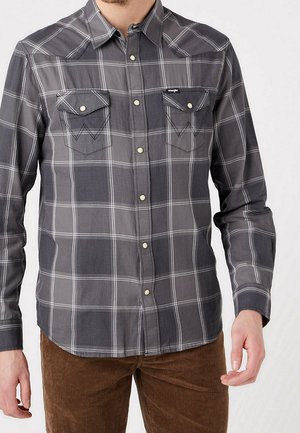 Mies, jolla on harmaa-musta ruutukuosinen nappillinen paita, jossa on kaksi etutaskua, sekä ruskeat corduroy-housut.