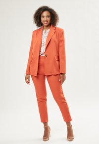 Oranges Anzugset mit einem Ein-Knopf-Blazer und schmal zulaufenden Hosen. Darunter eine leichte Bluse mit floralem Muster. Glattes Material mit strukturiertem Design.