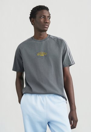 Grå bomulls-T-skjorte med korte ermer, med gul brodering og tre hvite striper langs skuldrene. Myk, avslappet passform.