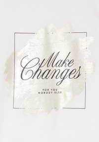 Biała bawełniana tkanina z metalicznym złotym nadrukiem z tekstem „Wprowadzaj zmiany” wewnątrz prostokątnej ramki, z dodatkowym tekstem poniżej.