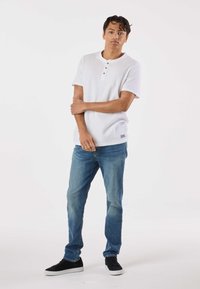 Valkoinen lyhythihainen henley-paita, jossa on kolmen napin läppä, yhdistetty vaalean sinisiin farkkuihin ja mustiin tennareihin, neutraalia taustaa vasten.