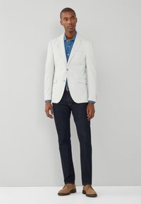 Hellgrauer Blazer über einem blauen Hemd, kombiniert mit einer dunklen Jeans und hellbraunen Schuhen. Der Blazer hat ein klassisches Design mit Reverskragen und Taschen.