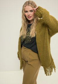 Olivgrüner Strickcardigan mit Fransenverzierung, getragen über einem schwarzen T-Shirt mit Grafik, kombiniert mit beigen, hochtaillierten Hosen.