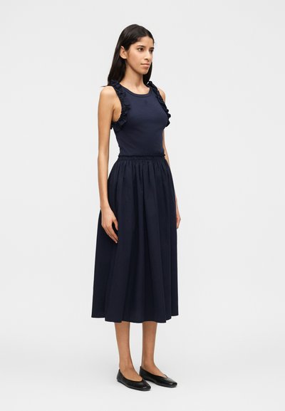 Γυναίκα με μπλε σκούρο αμάνικο τοπ με βολάν και αντίστοιχη μίντι φούστα με πιέτες, στέκεται φορώντας μαύρα φλατ παπούτσια σε απλό ανοιχτό γκρι φόντο.