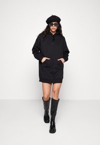 Schwarzes Hoodie-Kleid mit einer vorderen Tasche, Kordelzug-Kapuze und langen Ärmeln. Das Modell trägt schwarze knielange Stiefel und ein schwarzes Barett.