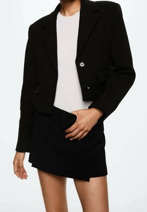 Woman wearing a black cropped blazer, white ribbed top, and asymmetrical black mini skirt, standing against a plain white background.