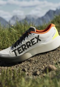Chaussure de trail Terrex Agravic 4 blanche et orange sur du gravier avec de l’herbe et des montagnes en arrière-plan.