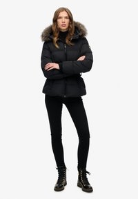 Giubbotto a sacco nero con cappuccio foderato in pelliccia, chiusura con zip e vita elasticizzata. Indossato con leggings neri e stivali neri con lacci.