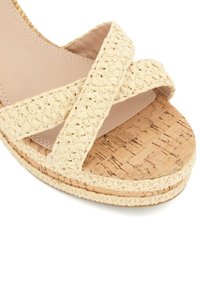 Sandale espadrille beige avec des brides croisées tissées et une plateforme en liège. Texture douce et finition naturelle, offrant un look décontracté.