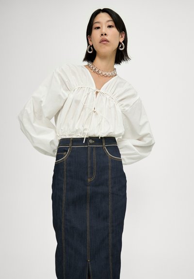 Chemisier blanc, froncé avec un col à cordon, associé à une jupe en denim bleu foncé taille haute avec des coutures contrastées et une fente à l'avant.