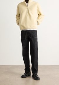 Pullover in pile beige con zip a un quarto e pantaloni in denim scuro. Il pullover presenta una superficie testurizzata; i pantaloni hanno cuciture a contrasto e tasche a toppa.