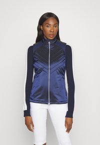 Modrá vycpaná vesta s vysokým límcem, zipem vpředu a kontrastními tmavými rukávy. Má design prošívání ve tvaru češky a hladký povrch.